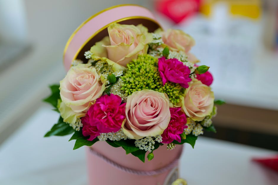 A close-up of a floral gift arrangement featuring pale pink roses, bright pink carnations, and small clusters of green buds, arranged in a pink round box with a decorative ribbon. The bouquet sits on 