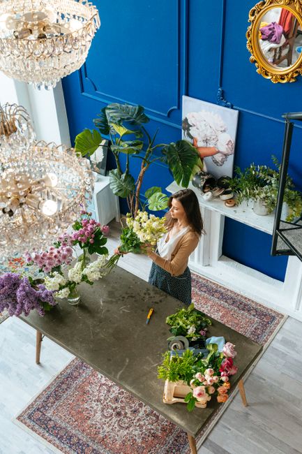 A woman in a beige top and a patterned skirt arranges a fresh floral bouquet inside a bright, modern flower studio with vibrant blue walls. The bouquet includes white lilies, green foliage, and pink r
